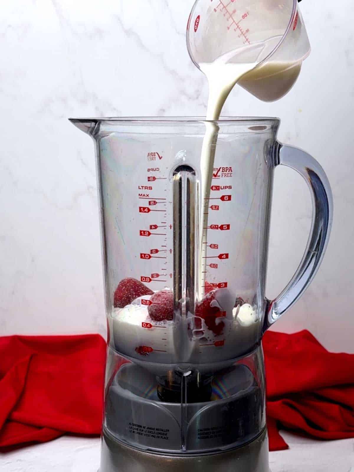 Adding milk to the blender with the frozen strawberries and strawberry fruit spread.