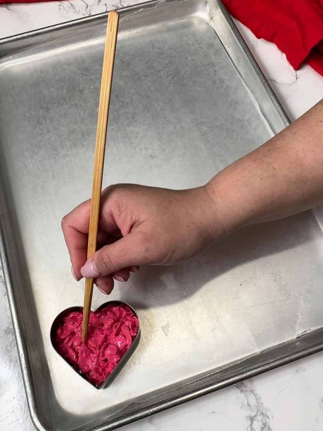 using chopstick to distribute bright pink corn flake mixture inside heart shaped cutter