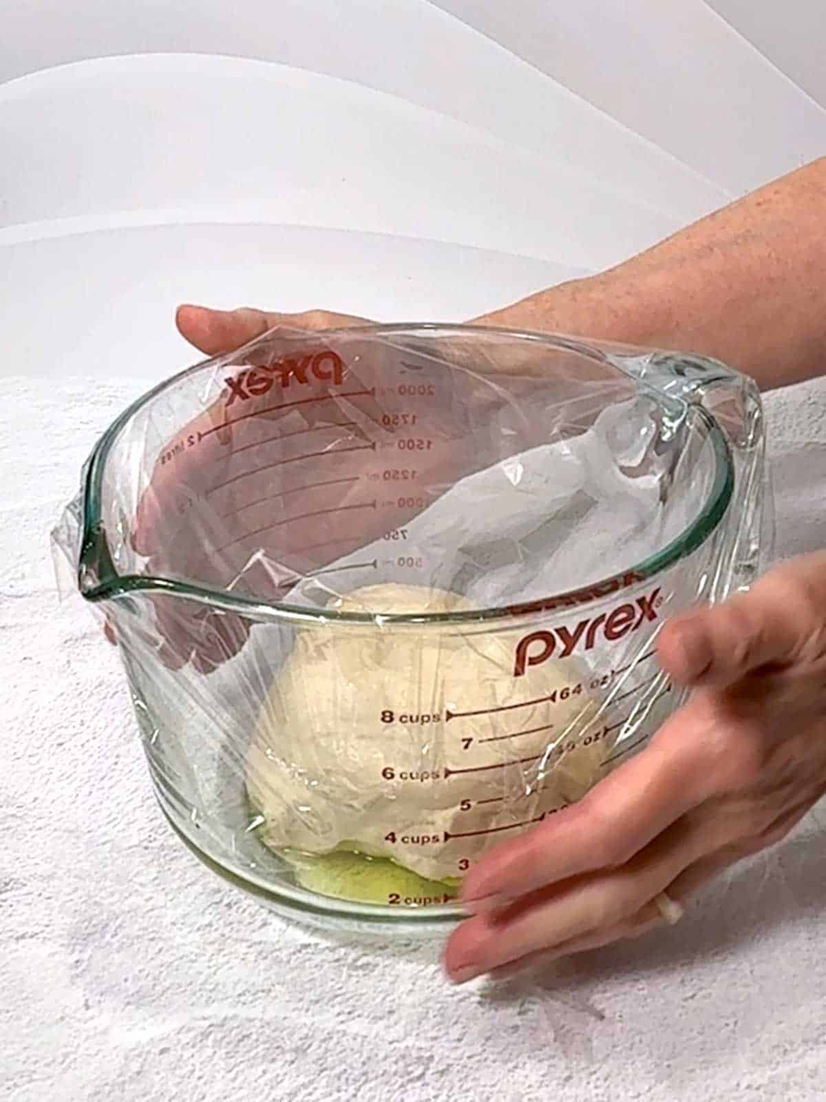 Covering the measuring cup with plastic wrap to allow the dough to proof.