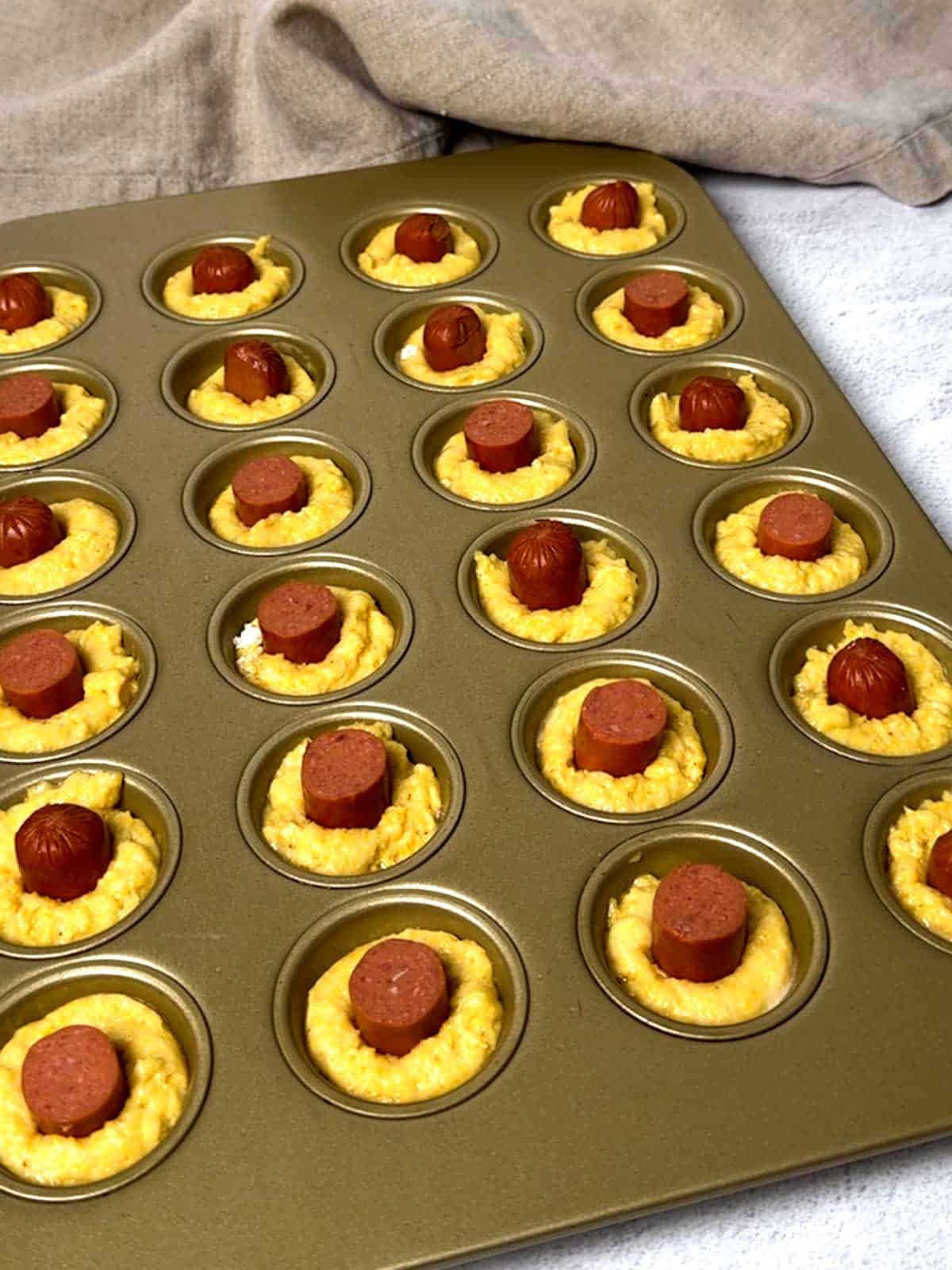 Mini muffin pan with cornbread batter and hot dog piece.